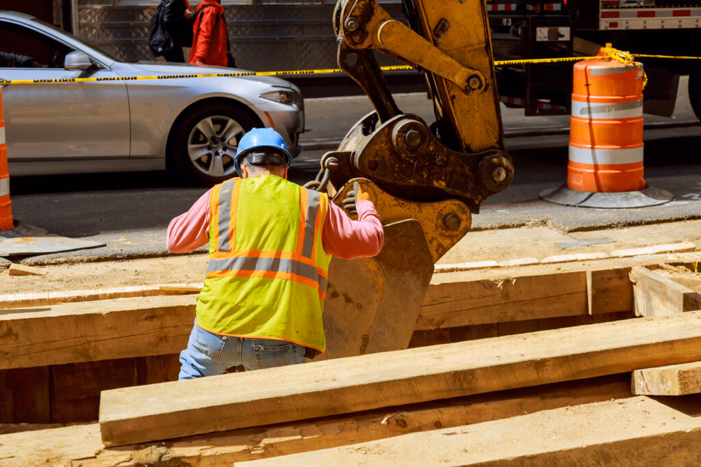 Worker guiding an excavator bucket in a shored trench on a city street