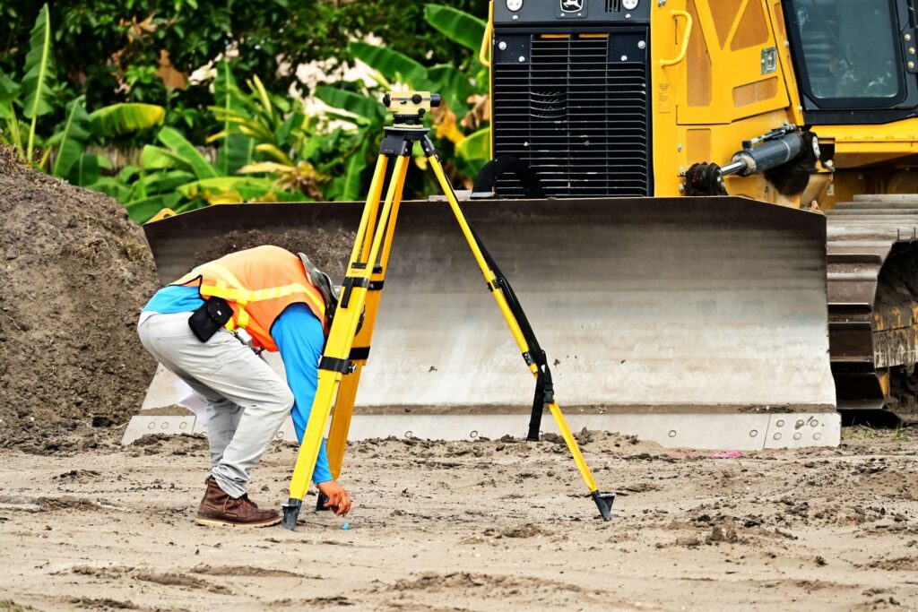 Surveyor in a safety vest setting up a total station in front of a bulldozer on a road construction site