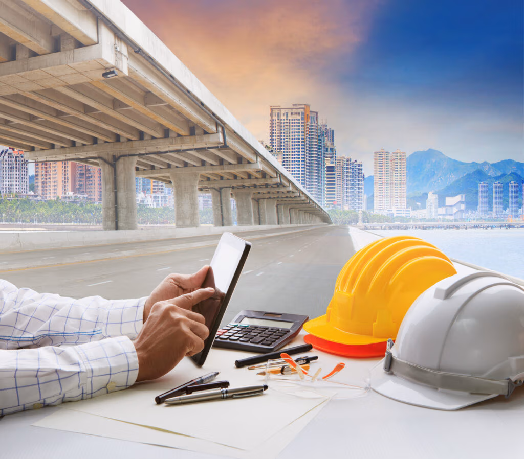 Desk with plans, a tablet and hard hats, with an overpass in the background