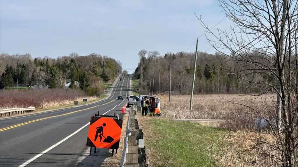 Field crew working on the shoulder of a rural highway behind a traffic sign